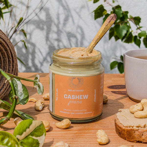 Organic cashew butter with a spoon, surrounded by cashews – plant prana