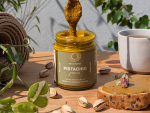 Organic nut butter – Jar of pistachio prana with a spoonful on a wooden surface with pistachios and a cup.