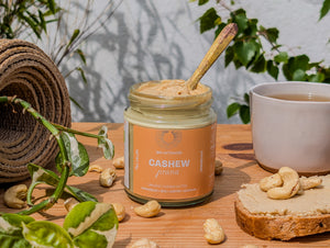 Activated Cashew Butter, an organic bio-activated cashew butter with a spoon, surrounded by cashews and a slice of bread on a wooden surface.